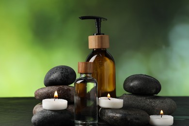 Photo of Spa composition with wet stones, cosmetic products and burning candles on dark table against blurred green background, closeup