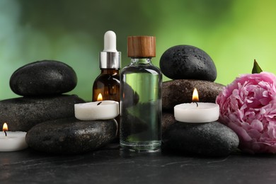 Photo of Spa composition with wet stones, cosmetic products and burning candles on dark textured table against blurred green background, closeup