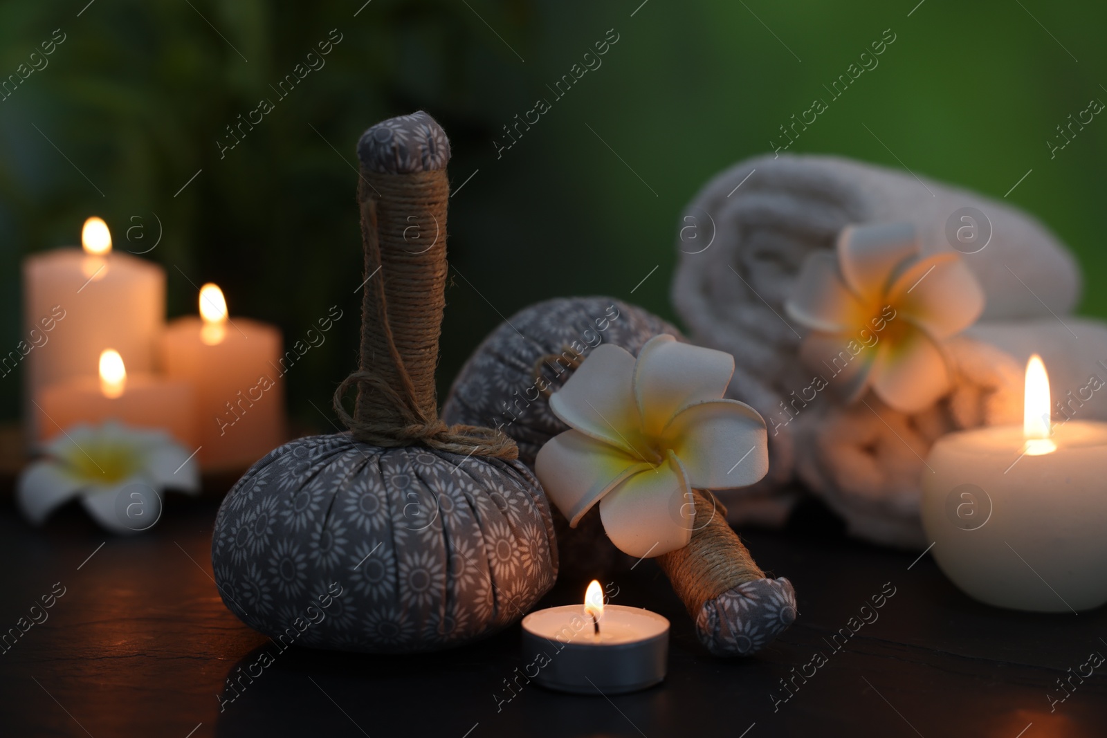 Spa composition with herbal bags, plumeria flowers and burning candles on dark table against blurred green background, closeup Photo of Spa composition with herbal bags, plumeria flowers and burning candles on dark table against blurred green background, closeup