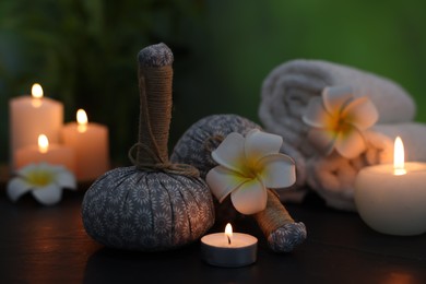 Photo of Spa composition with herbal bags, plumeria flowers and burning candles on dark table against blurred green background, closeup