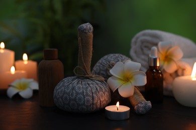 Photo of Spa composition with herbal bags, plumeria flowers and burning candles on dark table against blurred green background, closeup
