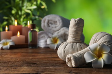 Photo of Spa composition with herbal bags, flowers and burning candles on wooden table against blurred green background, selective focus
