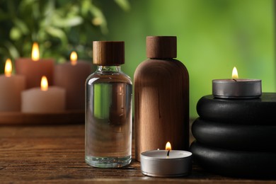 Photo of Spa composition with cosmetic products, stones and burning candles on wooden table against blurred green background, closeup