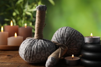 Photo of Spa composition with herbal bags, stones and burning candles on wooden table against blurred green background, closeup