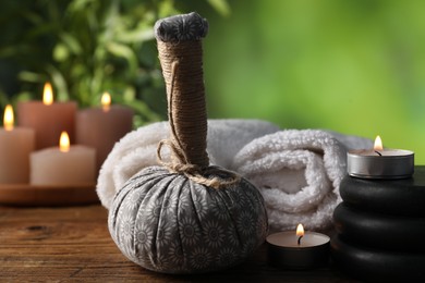 Photo of Spa composition with herbal bag, stones and burning candles on wooden table against blurred green background, closeup