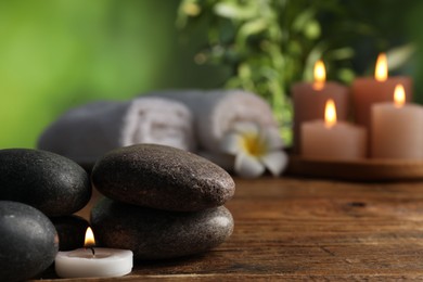 Photo of Spa composition with stones and burning candle on wooden table against blurred green background, closeup
