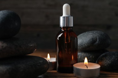 Photo of Spa composition with stones, dropper and burning candles on wooden table against black background, closeup