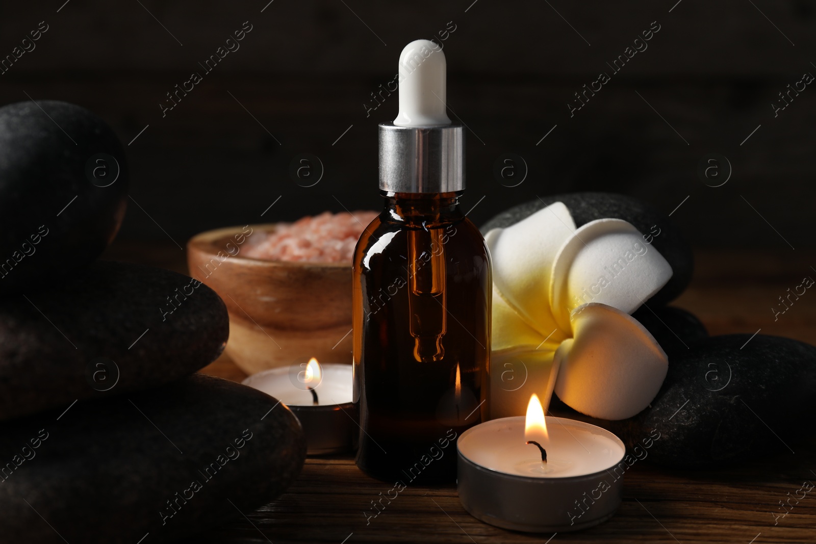Photo of Spa composition with stones, dropper and burning candles on wooden table against black background, closeup