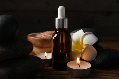 Photo of Spa composition with stones, dropper and burning candles on wooden table against black background, closeup