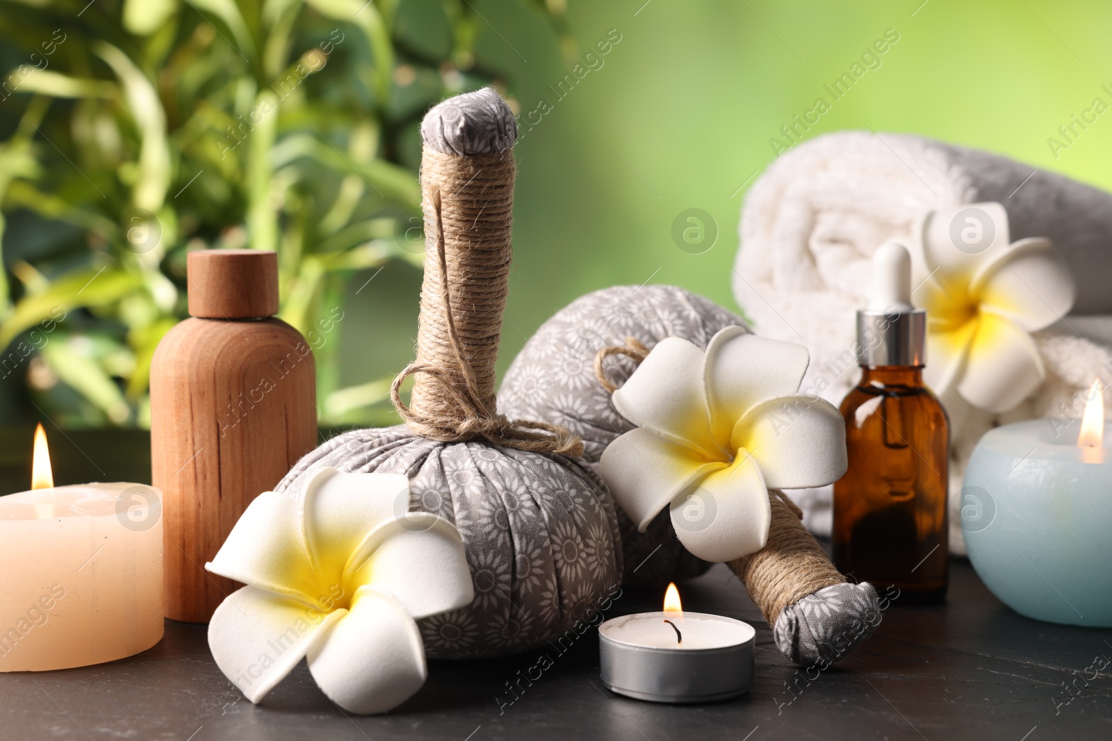 Spa composition with herbal bags, cosmetic products burning candles and plumeria flowers on grey textured table against blurred green background, closeup Photo of Spa composition with herbal bags, cosmetic products burning candles and plumeria flowers on grey textured table against blurred green background, closeup