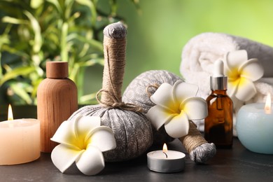 Photo of Spa composition with herbal bags, cosmetic products burning candles and plumeria flowers on grey textured table against blurred green background, closeup
