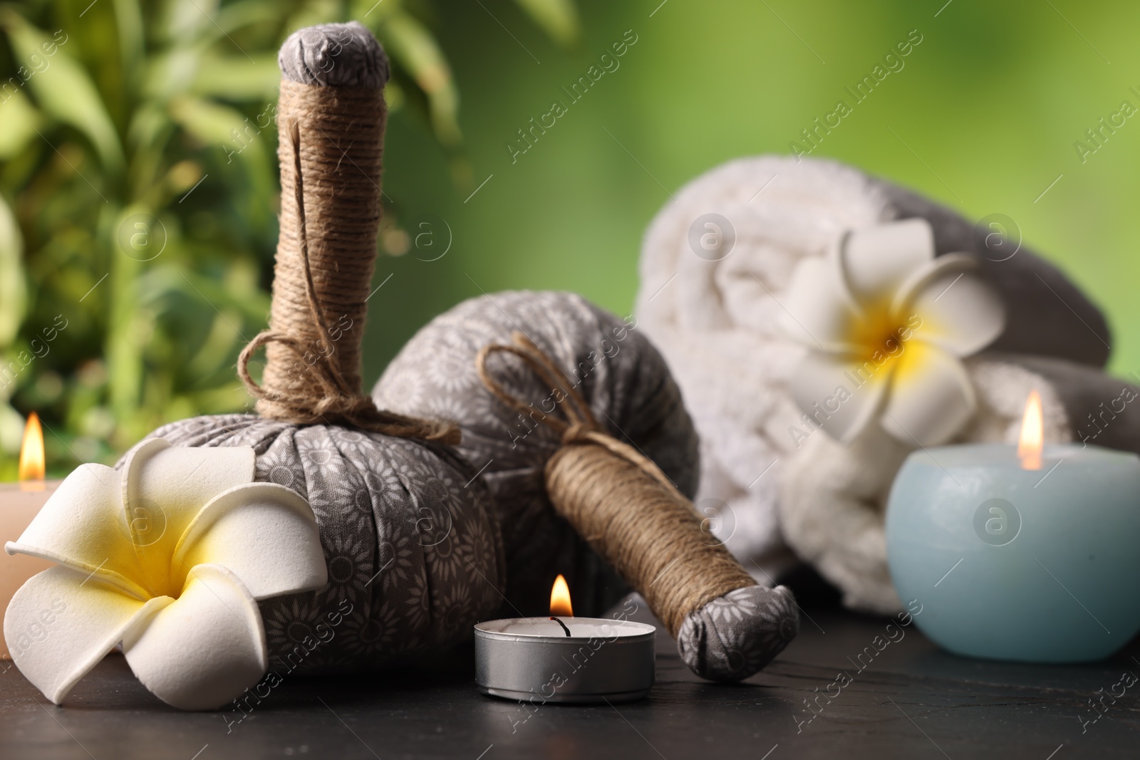 Spa composition with herbal bags, burning candles and plumeria flowers on grey textured table against blurred green background, closeup Photo of Spa composition with herbal bags, burning candles and plumeria flowers on grey textured table against blurred green background, closeup