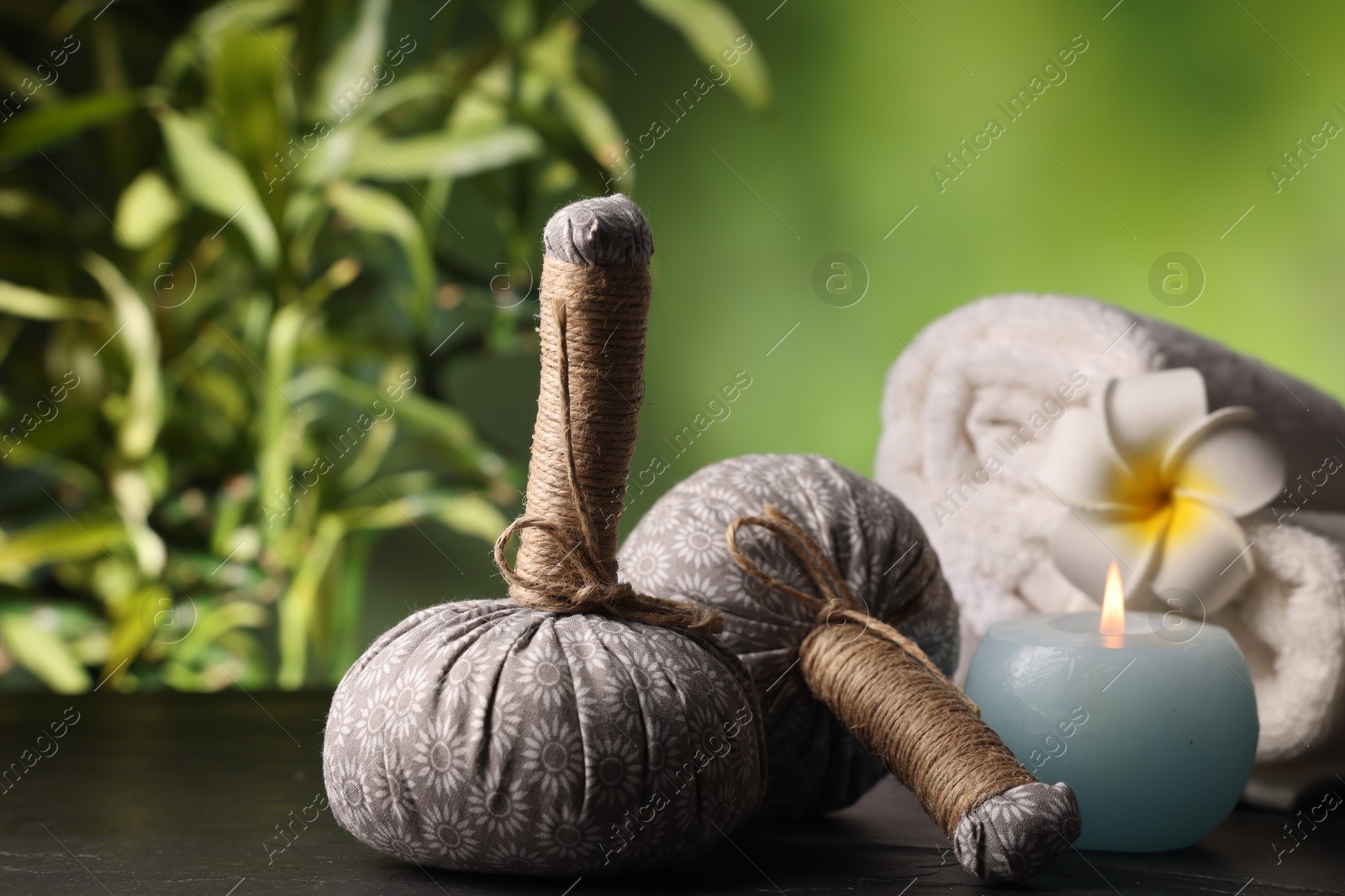 Spa composition with herbal bags and burning candle on grey textured table against blurred green background, closeup Photo of Spa composition with herbal bags and burning candle on grey textured table against blurred green background, closeup