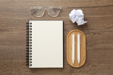 Photo of Notebook, pens, crumpled paper and glasses on wooden table, flat lay