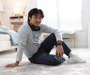 Photo of Handsome Asian man with headphones and smartphone on floor at home