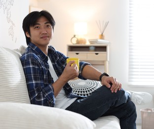 Photo of Handsome Asian man with cup of hot drink on sofa at home