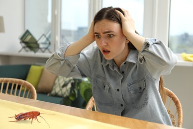 Image of Emotional young woman looking fearfully at cockroach on table indoors