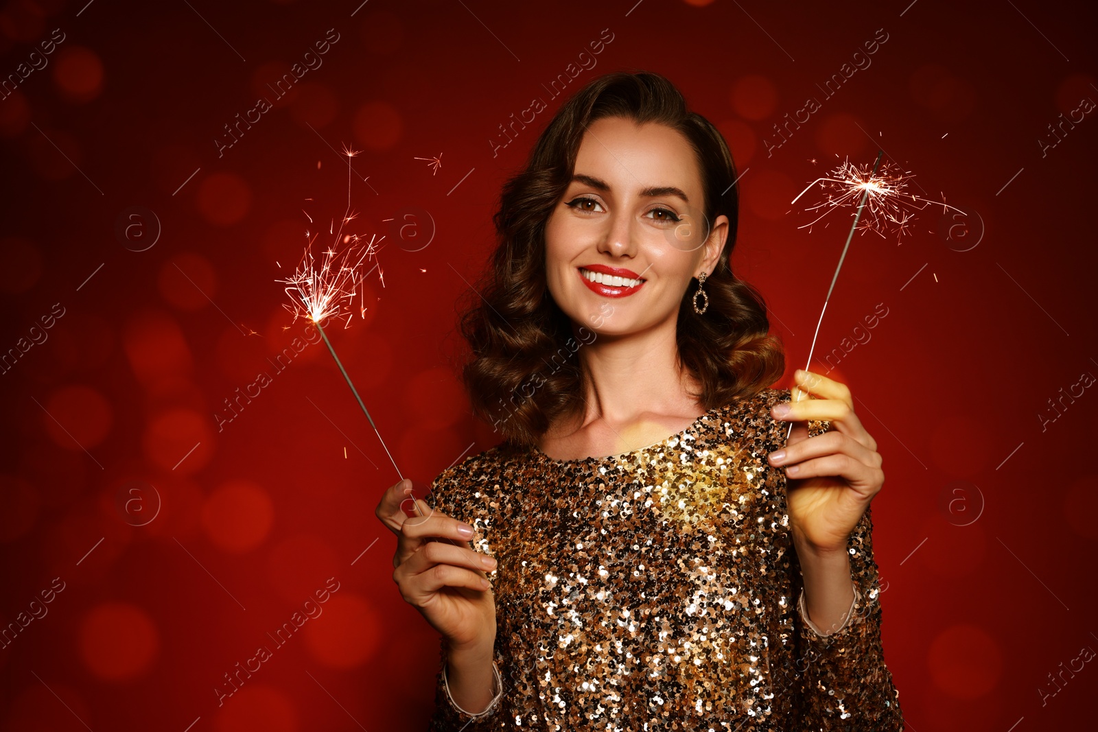 Smiling woman with sparklers celebrating Christmas on red background with blurred lights, bokeh effect Image of Smiling woman with sparklers celebrating Christmas on red background with blurred lights, bokeh effect