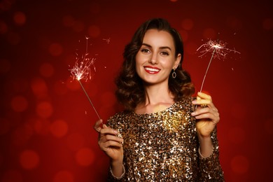 Image of Smiling woman with sparklers celebrating Christmas on red background with blurred lights, bokeh effect