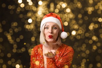 Image of Woman with perfect makeup in Santa hat blowing confetti on black background, bokeh effect. Christmas celebration