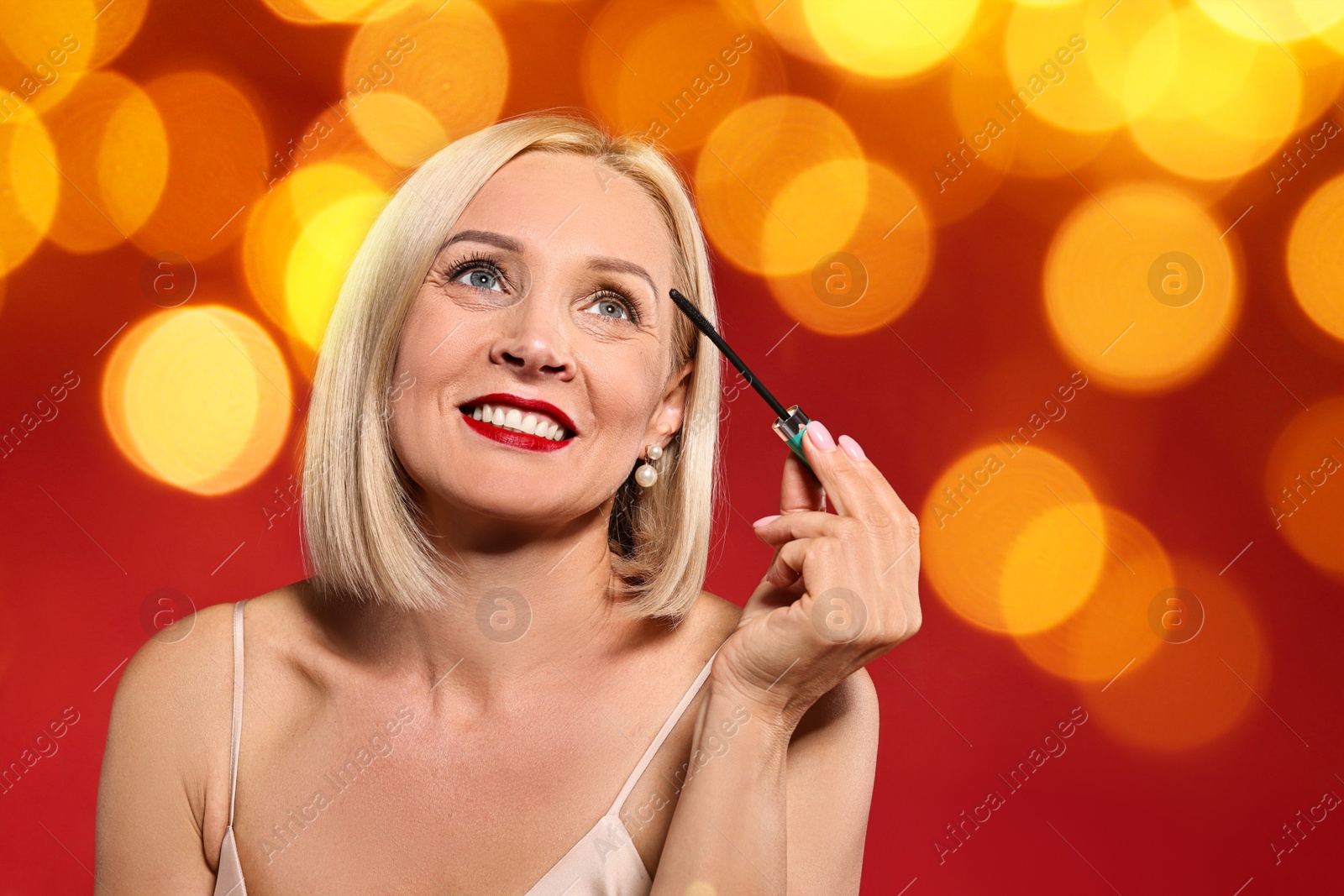 Smiling woman with perfect makeup applying mascara on red background, blurred lights. Bokeh effect Image of Smiling woman with perfect makeup applying mascara on red background, blurred lights. Bokeh effect