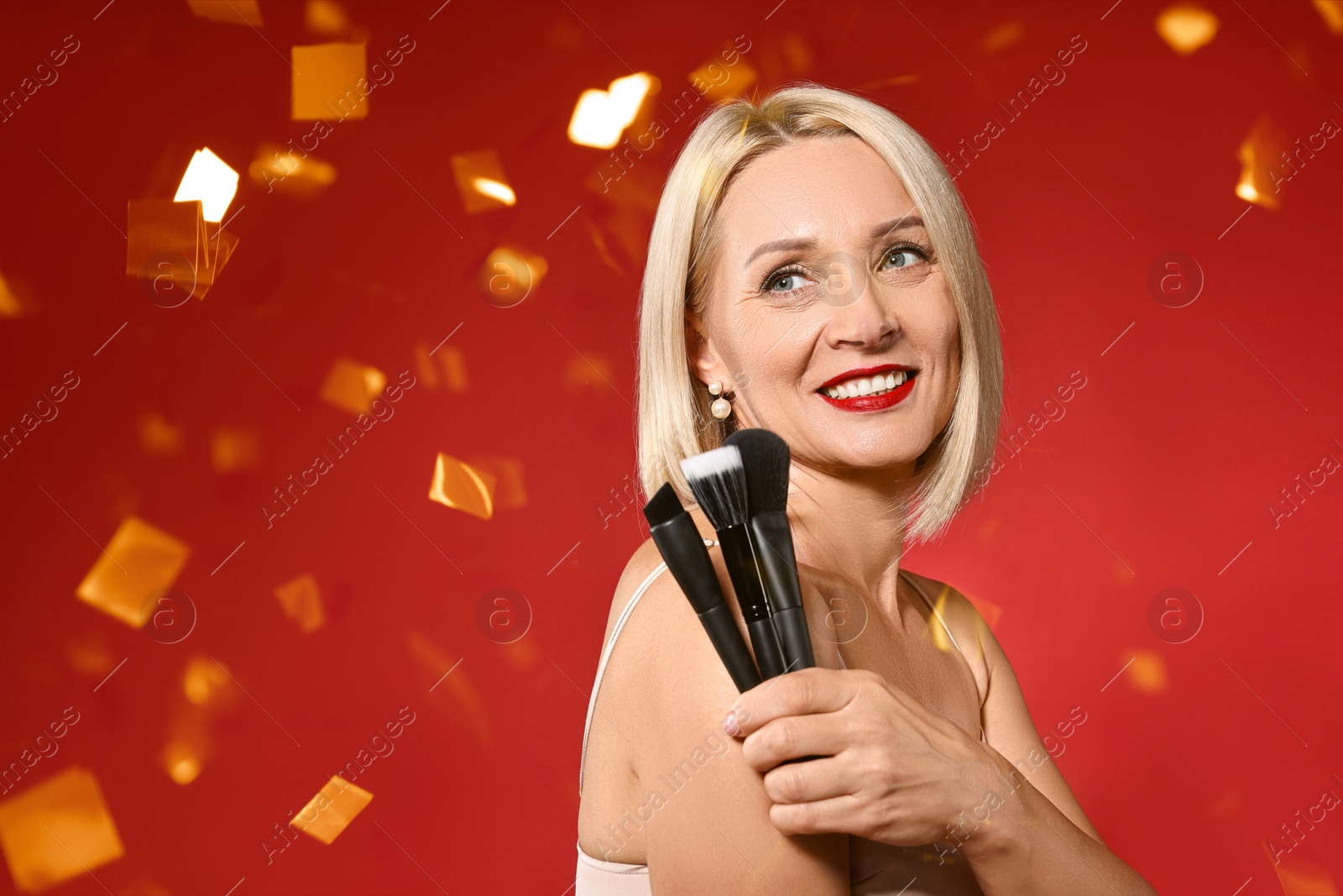 Smiling woman with different makeup brushes under falling confetti on red background Image of Smiling woman with different makeup brushes under falling confetti on red background