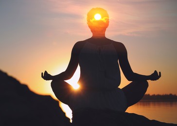 Image of Mental health. Woman meditating near river at sunrise. Sun shining in her head