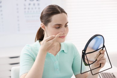 Photo of Young woman putting on contact lenses near mirror in ophthalmologist office
