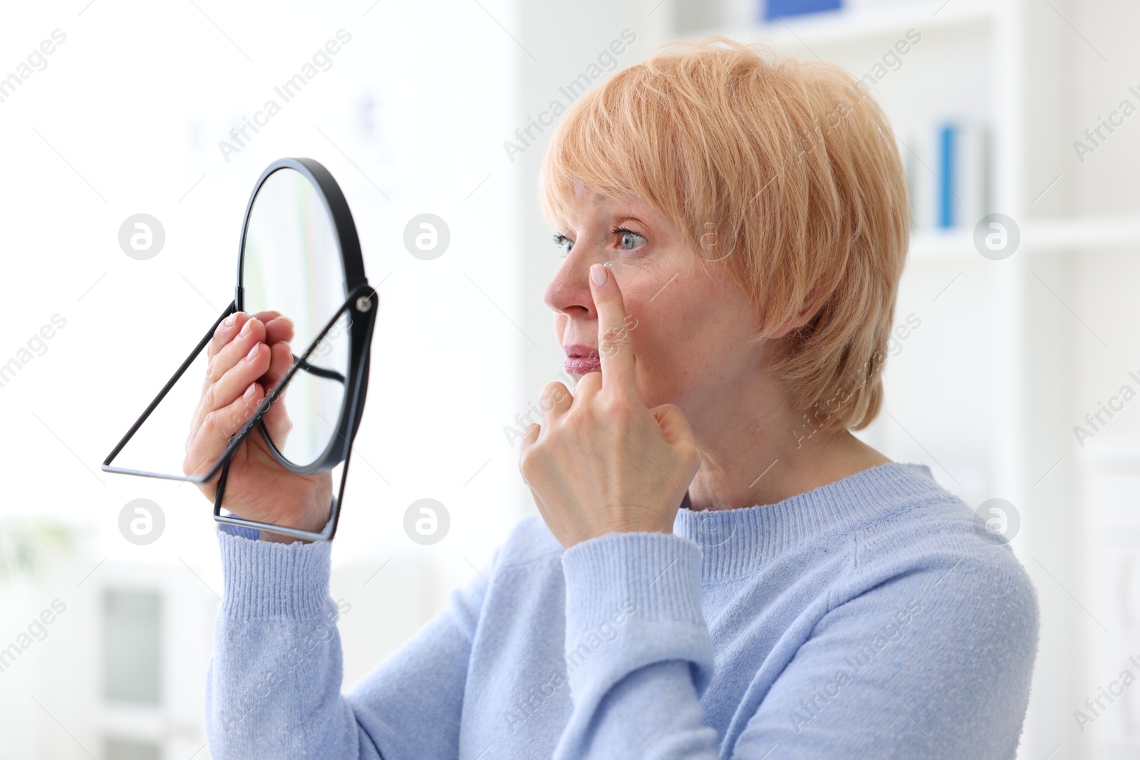 Photo of Senior woman putting on contact lenses near mirror in ophthalmologist office