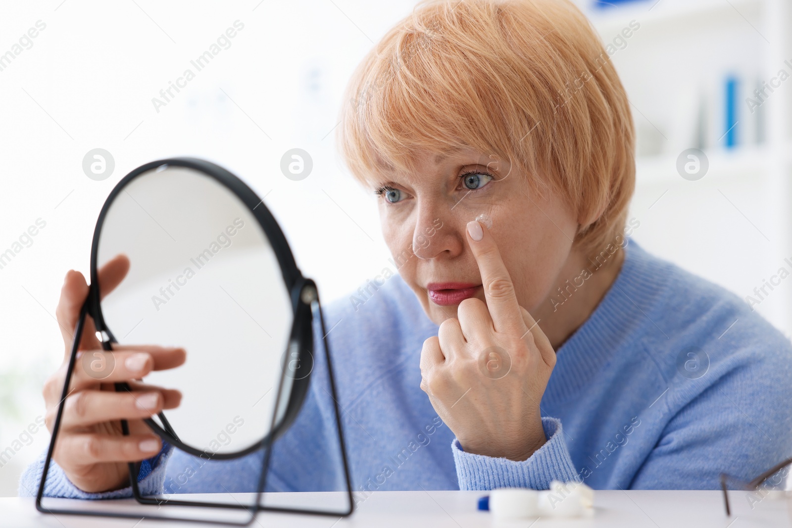 Senior woman putting on contact lenses at table near mirror in ophthalmologist office Photo of Senior woman putting on contact lenses at table near mirror in ophthalmologist office