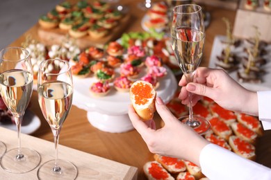 Photo of Buffet menu. Woman taking glass of sparkling wine and caviar sandwich from table, closeup