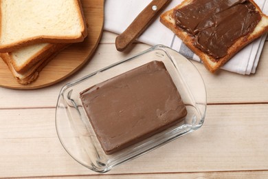 Photo of Block of tasty chocolate butter in keeper and slices of bread on light wooden table, flat lay