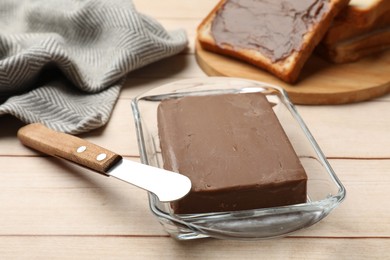 Photo of Block of tasty chocolate butter in glass keeper and knife on light wooden table, closeup