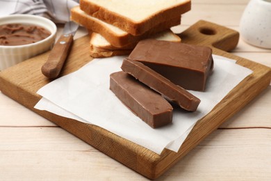 Photo of Pieces of tasty chocolate butter and slices of bread on light wooden table, closeup