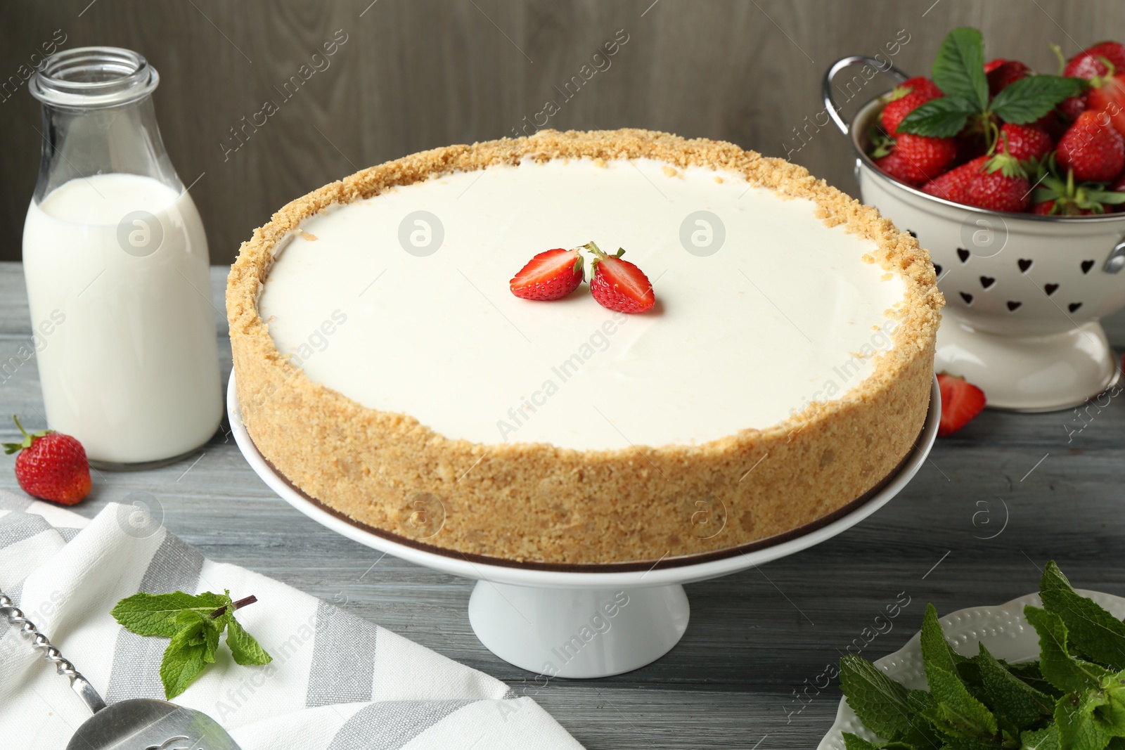 Photo of One tasty cheesecake, strawberries, mint and milk on grey table against wooden background, closeup