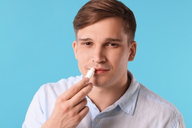 Photo of Young man applying lip balm onto his lips on light blue background