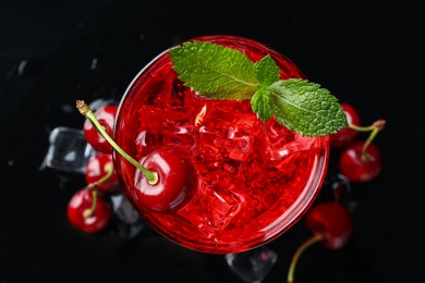 Photo of Tasty cherry soda with ice cubes, berries and mint in glass on black background, top view