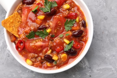 Photo of Tasty chili con carne in bowl and nachos on grey textured table, top view