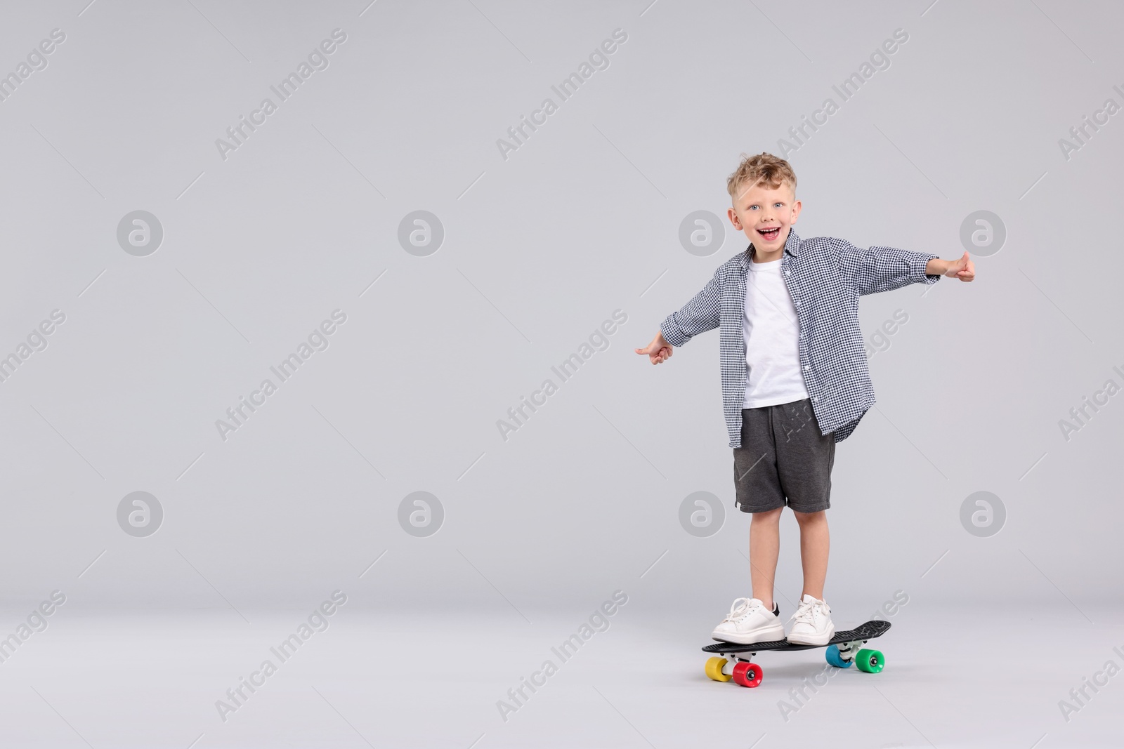Cheerful little boy standing on penny board and showing thumbs up against gray background, space for text Photo of Cheerful little boy standing on penny board and showing thumbs up against gray background, space for text