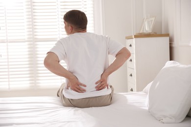 Photo of Young man suffering from back pain on bed with soft mattress at home, back view