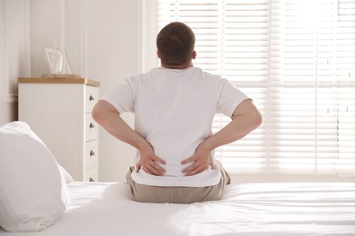 Photo of Young man suffering from back pain on bed with soft mattress at home, back view