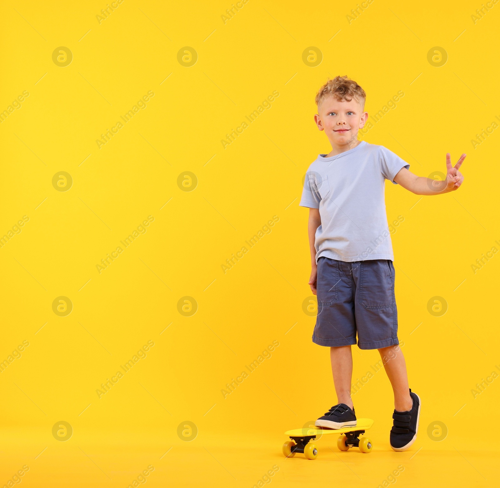 Little boy with penny board showing V-sign on yellow background, space for text Photo of Little boy with penny board showing V-sign on yellow background, space for text
