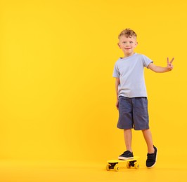 Photo of Little boy with penny board showing V-sign on yellow background, space for text