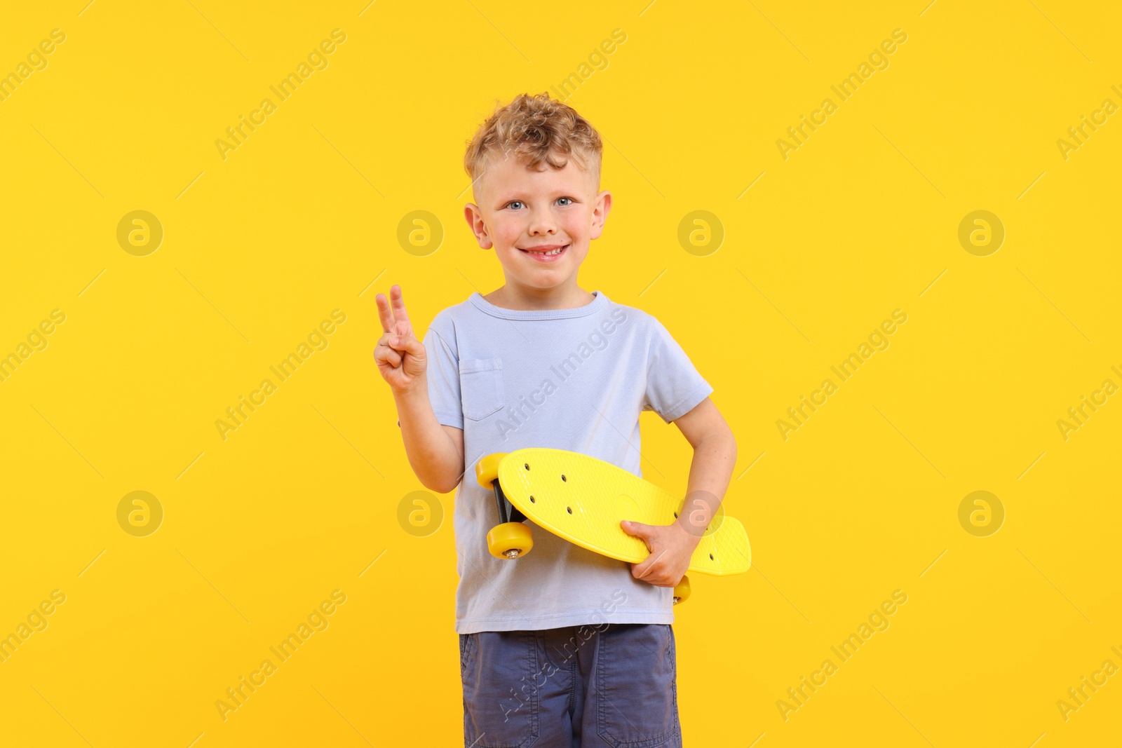 Little boy with penny board showing V-sign on yellow background Photo of Little boy with penny board showing V-sign on yellow background