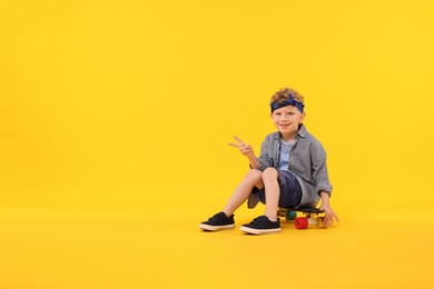 Photo of Little boy sitting on penny board and showing V-sign against yellow background, space for text