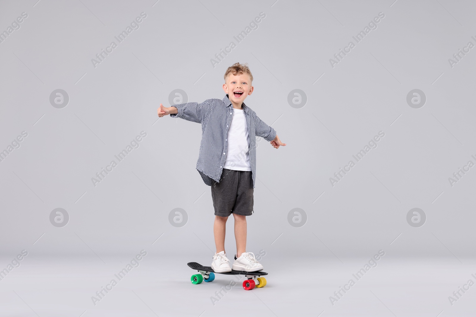Cheerful little boy standing on penny board and showing thumbs up against gray background Photo of Cheerful little boy standing on penny board and showing thumbs up against gray background