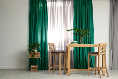Image of Dining table and chairs near window with green curtains in room. Interior design