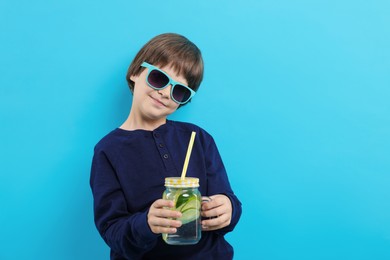 Photo of Cute little boy in sunglasses with mason jar of lemonade on light blue background. Refreshing drink