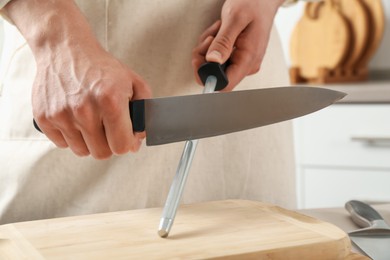 Photo of Man sharpening knife with sharpener at table indoors, closeup
