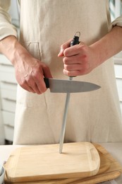Photo of Man sharpening knife with sharpener at table indoors, closeup
